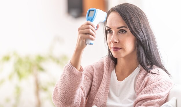 Woman self-measuring temperature with an infrared digital thermometer on her forehead.