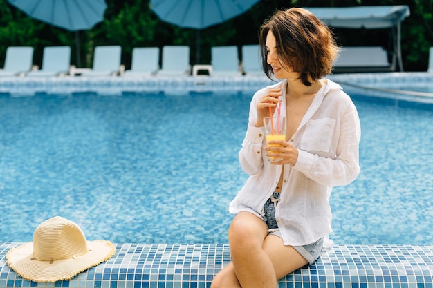 A woman in a shirt drinks freshly squeezed juice from a glass while sitting on the edge of the swimming pool during her summer vacation or relaxing by the water Latina girl heals with juice