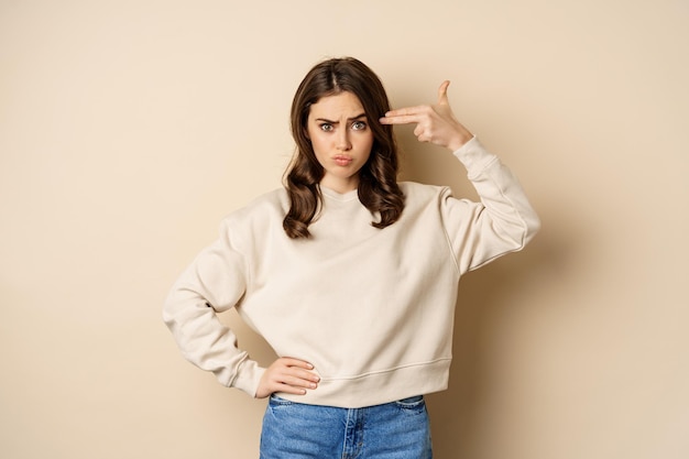 Woman showing finger pistol gesture over her head, being bothered and annoyed, standing over beige background.