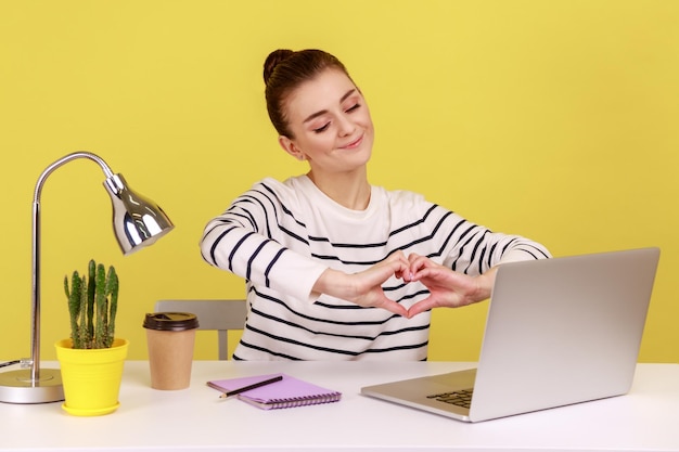 Woman showing heart gesture with hands to laptop screen sitting at workplace with laptop