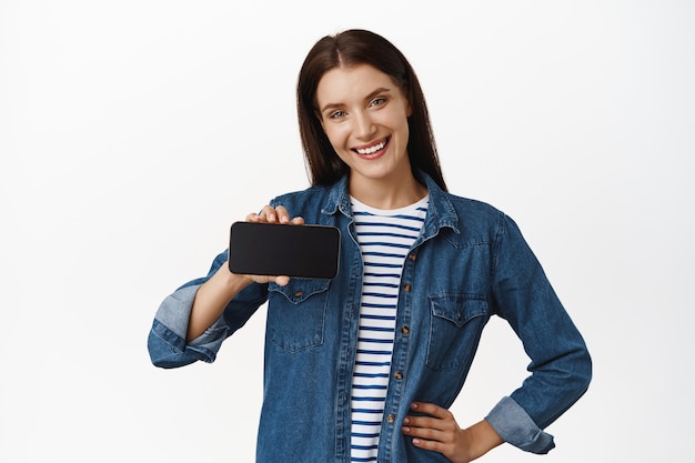 woman showing smartphone empty display, app interface on horizontal screen, smiling satisfied, recommending application on mobile phone on white.