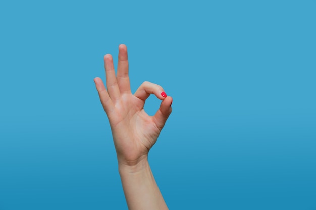 Woman shows ok gesture. Female hand with red gel polish manicure on a blue wall Copy space