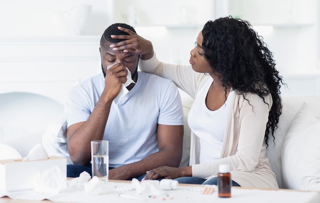 A woman sits beside her sick partner holding his forehead and offering comfort as he blows his nose