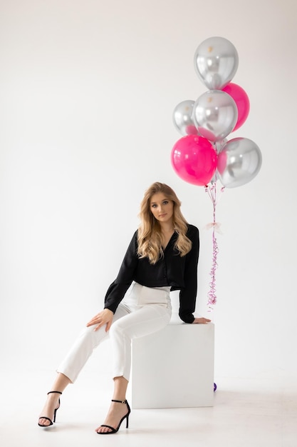 A woman sits on a box with a bunch of pink and silver balloons.