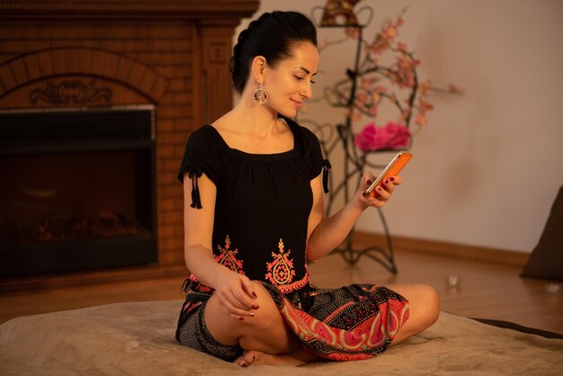 A woman sits on a carpet in front of a fireplace, using a phone.