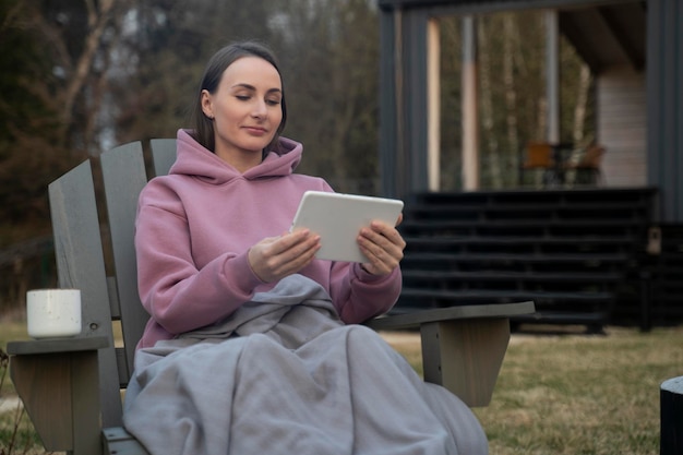 Woman sits on a chair by the fire and uses a digital tablet relaxing in the backyard during sunset t