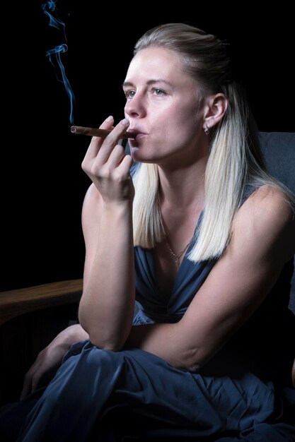 Photo woman sits in a chair and smokes a cigarette, holding it in her mouth, isolated on a black background