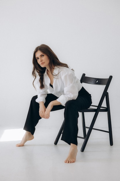 A woman sits on a chair in a white room, wearing black pants and a white shirt.