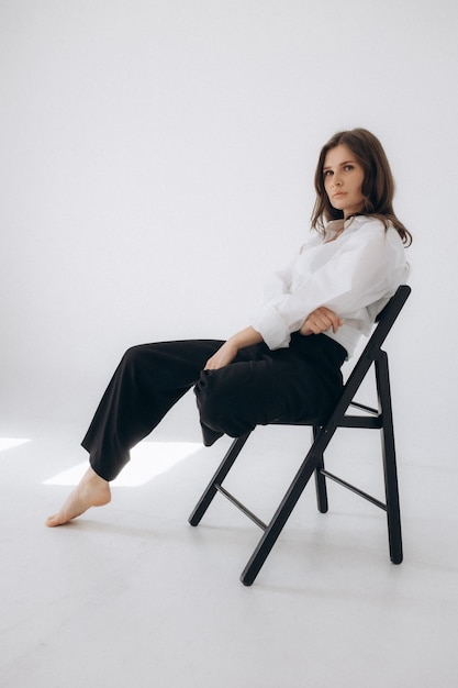 A woman sits on a chair in a white shirt and black pants.