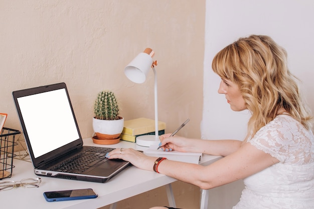 A woman sits at a laptop with a phone and writes in a notebook