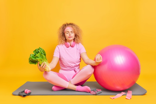 woman sits in lotus pose tries to relax meditates after fitness training practices yoga holds green vegetable sits on mat near fitball sport equipment 