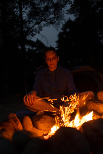 A woman sits at night by a campfire and drinks tea against the background of a fire on a hike