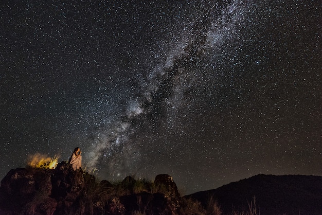 Woman sits on stone ledge looking at the night sky stars with milky way background