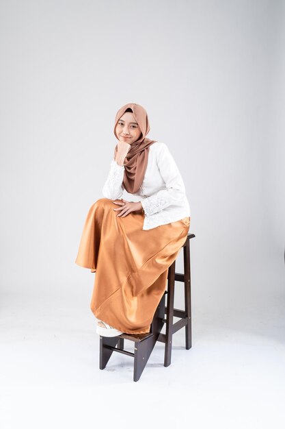 A woman sits on a stool wearing a hijab and a white shirt.