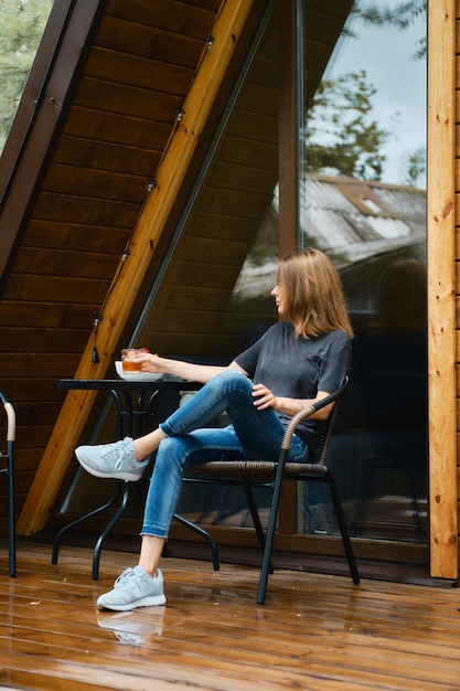 Woman sits in wicker chair near the table at wooden terrace