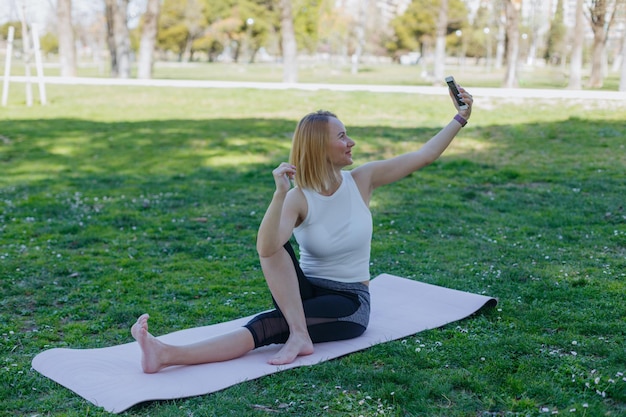 A woman sits on a yoga mat in a park, taking a self portrait.