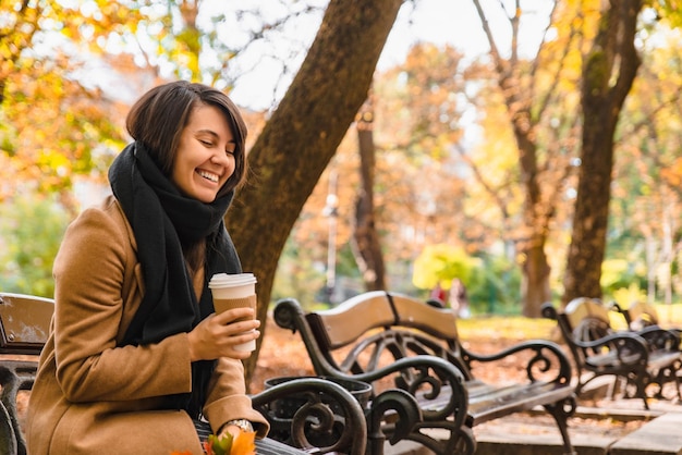 Woman sitting on the bench at autumn city park drinking coffee