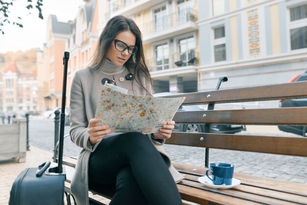 Woman sitting on bench with travel suitcase, reading city map