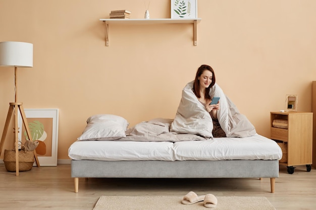 Woman sitting on comfortable bed at home wrapped in blanket with phone