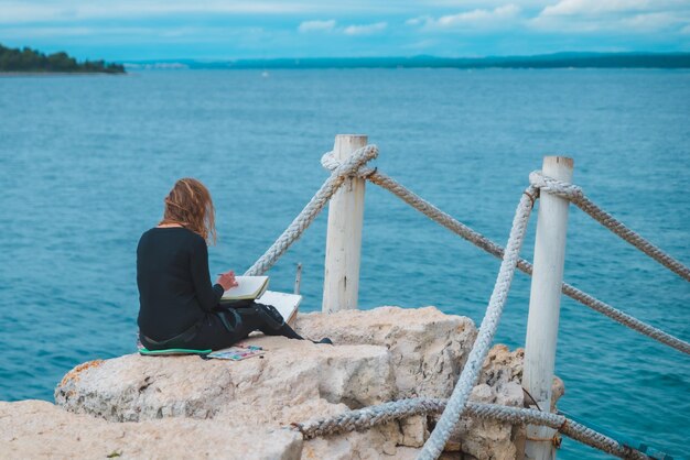 Woman sitting at the edge drawing picture of seascape suspension bridge