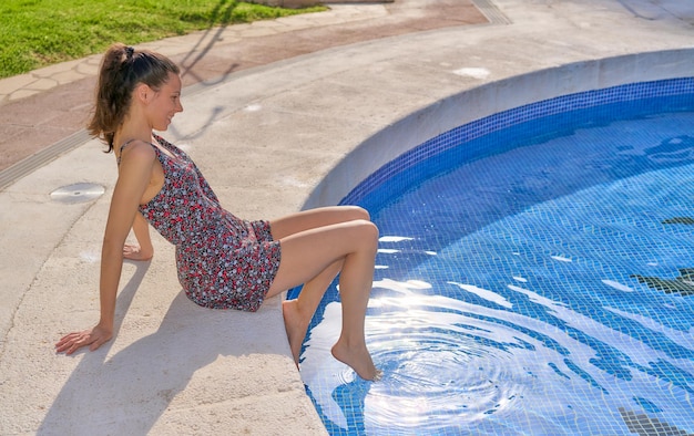 Woman sitting on the edge of the pool sunbathing in a dress with her feet in the water