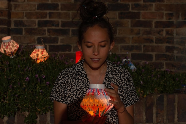 Photo woman sitting on the ground holding a paper lantern surrounded by several lanterns celebrating the day of the candles in colombia