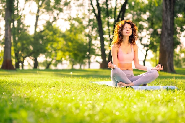 Woman sitting in lotus position with closed eyes on yoga mat in the park copy space