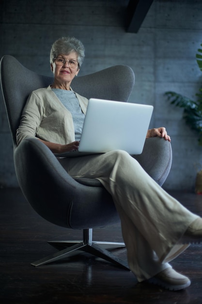 Woman sitting on modern chair near the window in light cozy room at home She is working on laptop in relaxing atmosphere