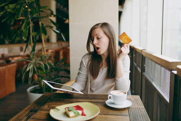 Woman sitting near big window in coffee shop at table with credit card, cup of coffee cake relaxing in restaurant during free time. Female working on tablet pc computer rest in cafe. Lifestyle concept