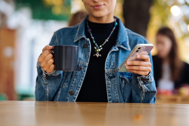 Woman sitting outdoors cafe drinking beverage holding in hand smartphone