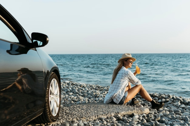 Woman sitting on rocks with juice looking at sea near car