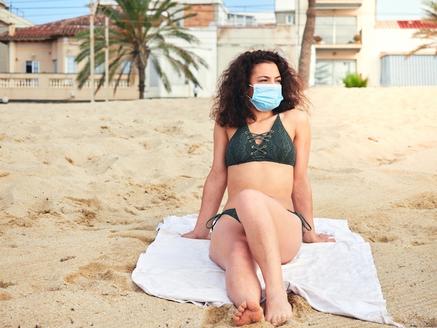 Woman sitting on the sand on the beach sunbathing with a medical mask