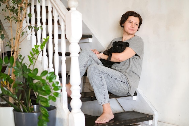 Woman sitting on stairs, using smartphone with black griffon or pug on her lap