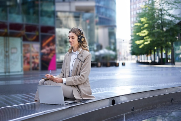 Woman sitting on a street with laptop and headphones plugged in taking notes corporate worker attend