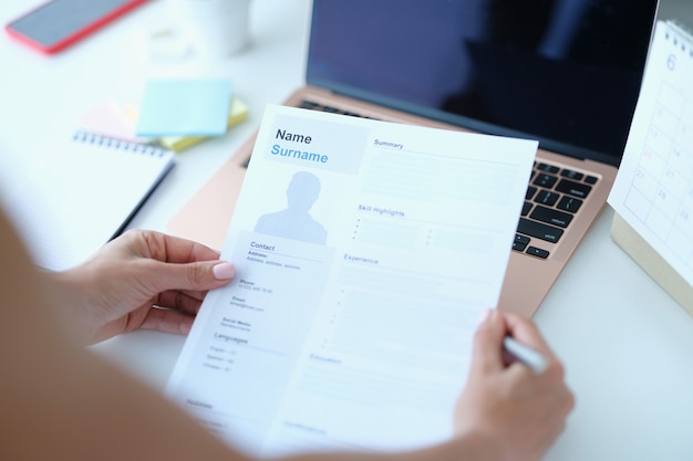 Photo woman sitting at table in front of laptop and holding paper with resume for employment in her hands