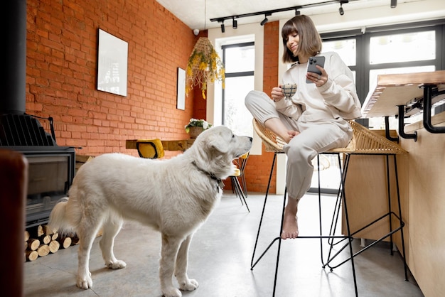 Woman sitting with a coffee cup and phone at bar table with her dog in sunny living room