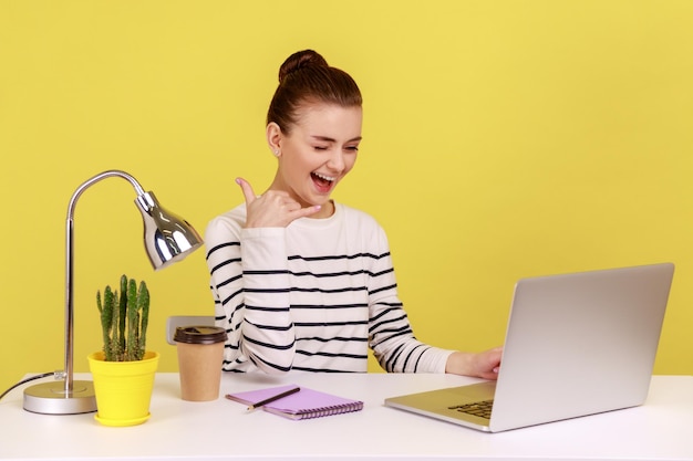 Woman sitting at workplace keeping hand shaped like telephone near head looking at laptop screen