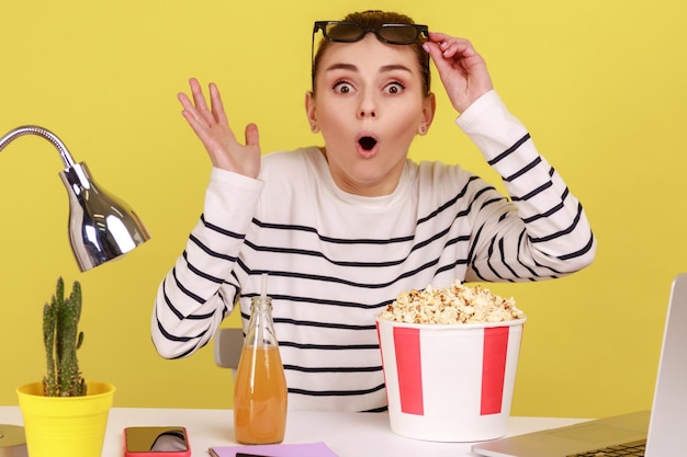 Woman sitting at workplace with popcorn and beverage raising glasses and watching shocking moment