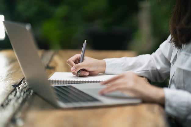 Woman in smart casual wear writing on notebook and working on laptop while sitting in creative office or cafe young girl working with laptop on the wood table in the cafe