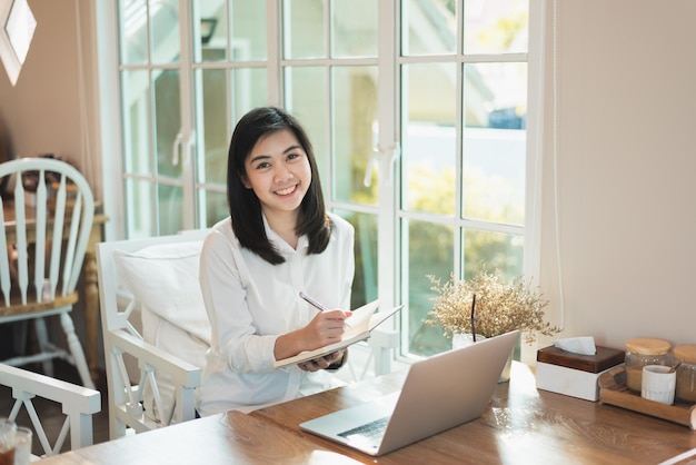 Woman in smart casual wear writing on notebook and working on laptop while sitting in creative office or cafe young girl working with laptop on the wood table in the cafe