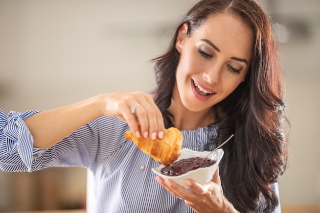 Woman smiles as she enjoys breakfast with croissant and jam.