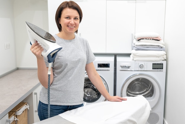 Woman smiling while standing near ironing board in laundry room with washing machine 