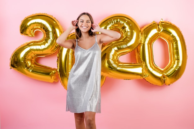 Woman smiling with 2020 new year balloons wearing dress isolated over pink