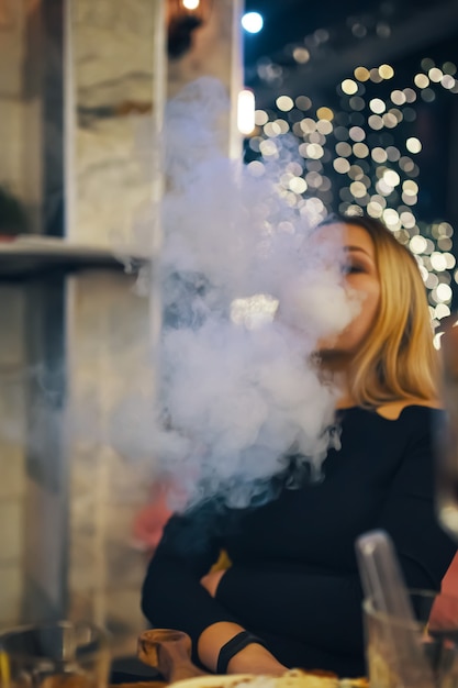 Woman smokes hookah in lounge bar female in black dress is resting at cafe table bright lights on ba...