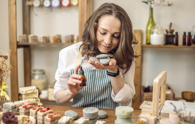 Woman soap maker in a workshop holds pieces of natural soap and a bottle of oil in her hands