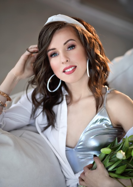 woman on the sofa in a white shirt and blue jeans with a bouquet of lilies of the valley. Portrait of a young woman in a white interior.