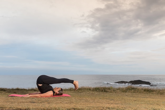 Woman in sportswear doing pilates outdoors on a pink mat with the sea