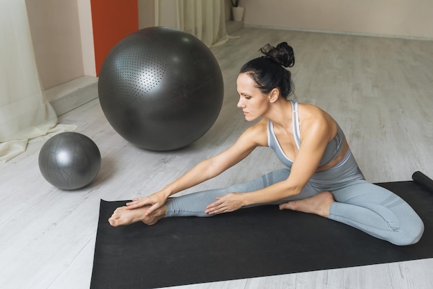 A woman in sportswear performs an ankle and back muscle stretching exercise while sitting on a mat