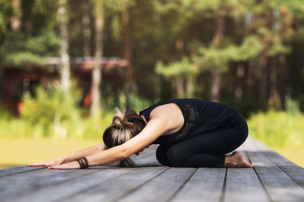 Woman in sportswear sitting on a wooden bridge practices yoga asanas performing a childs pose