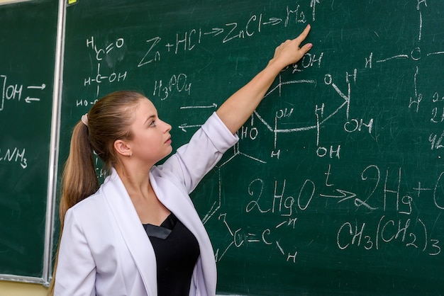 Woman standing in chemist's classroom near board and pointing on it